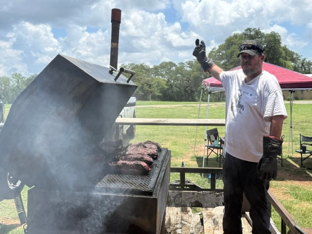 A Johnson City Lodge pitmaster tending briskets over a smoky outdoor pit.
