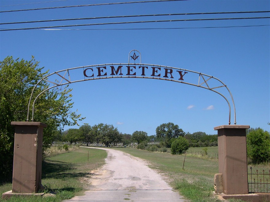 The wrought-iron entrance arch to the Johnson City Masonic Cemetery, topped by the Square and Compasses of Freemasonry, with the gravel road leading into the cemetery beyond.
