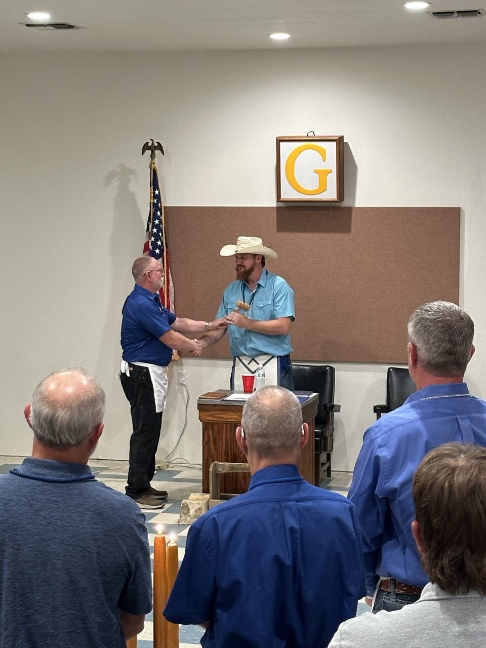 The passing of the gavel between officers in the East — both brothers wearing Masonic aprons, the framed letter G visible on the wall behind them.
