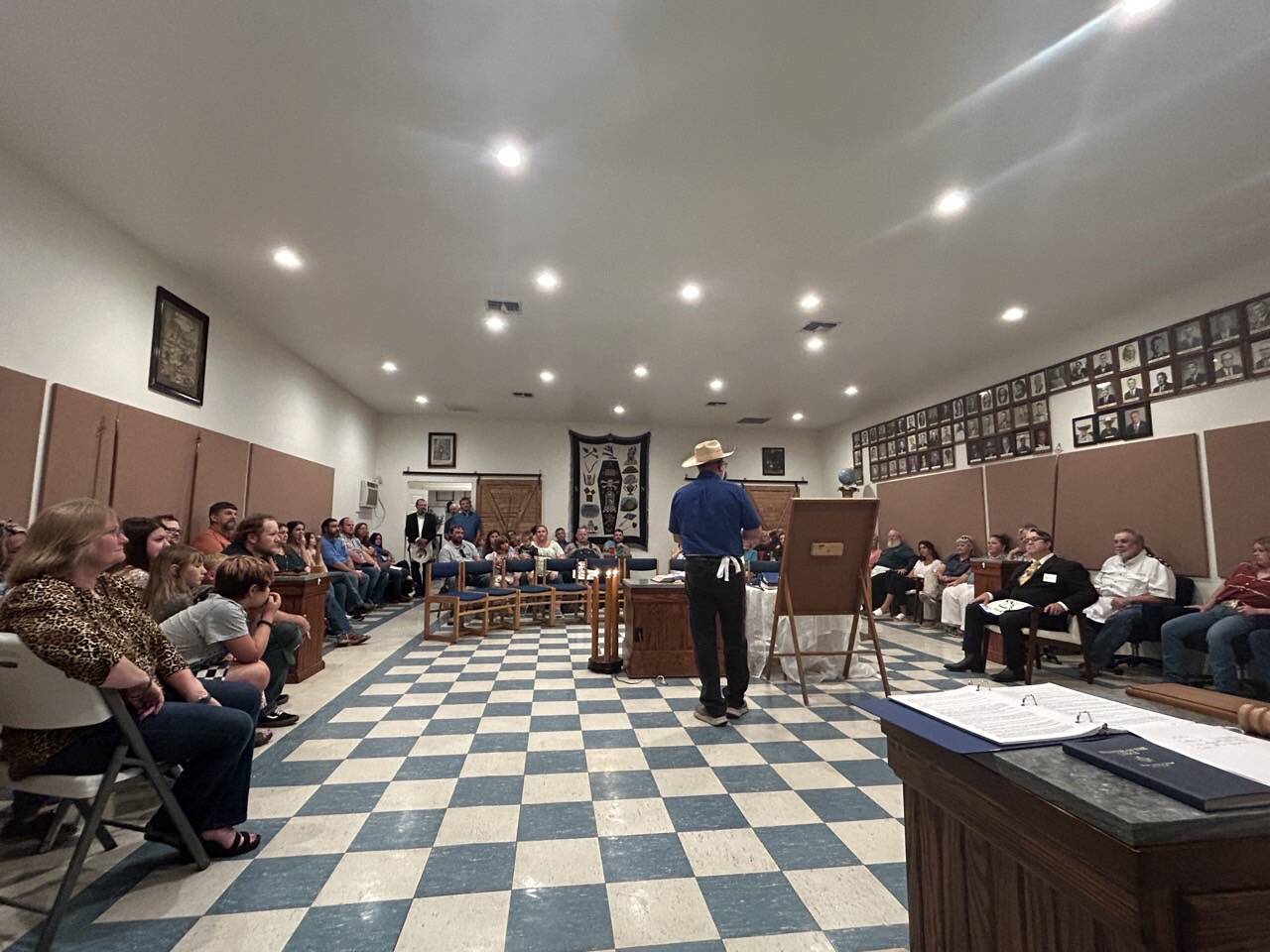 The interior of the lodge room with its blue-and-white checkered floor, past masters portraits along the walls, and a speaker addressing seated guests.