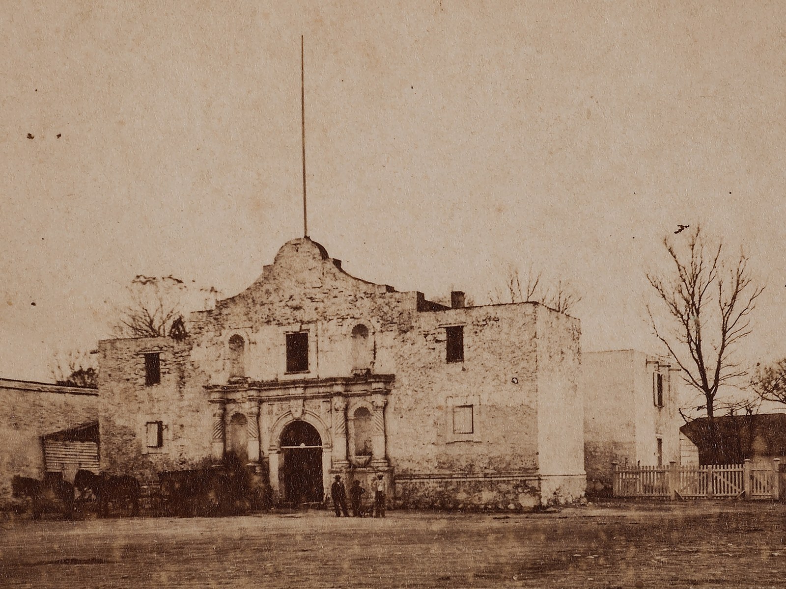 Sepia 1860s photograph of the Alamo church in San Antonio, with its carved stone doorway and curved gable, a tall flagstaff rising above the roofline, and three men standing near the entrance.