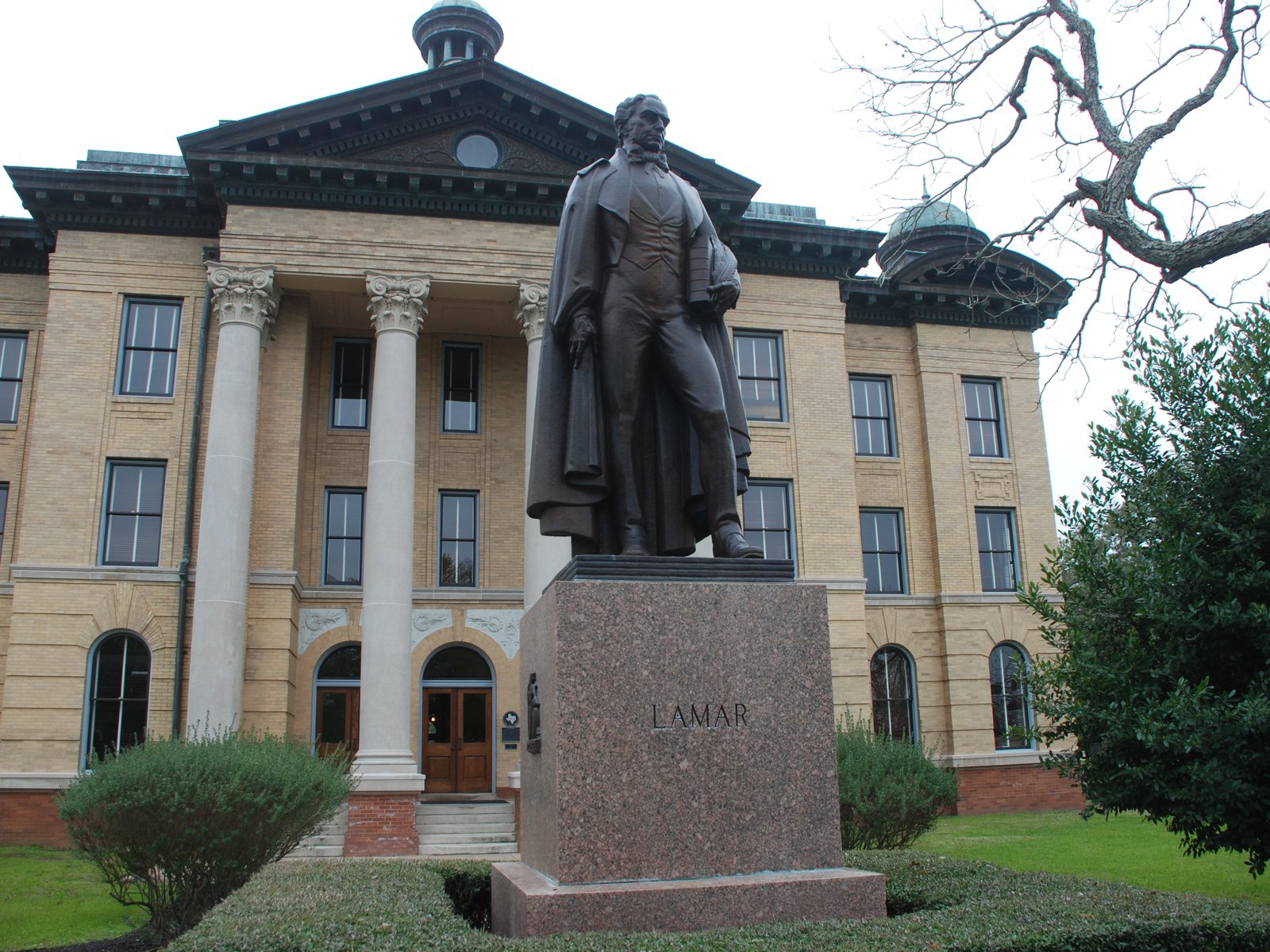 Statue of Mirabeau B. Lamar in a long flowing cape, holding a document at his side, standing on a red granite base carved with the name LAMAR before the columned facade of the Fort Bend County Courthouse in Richmond, Texas.